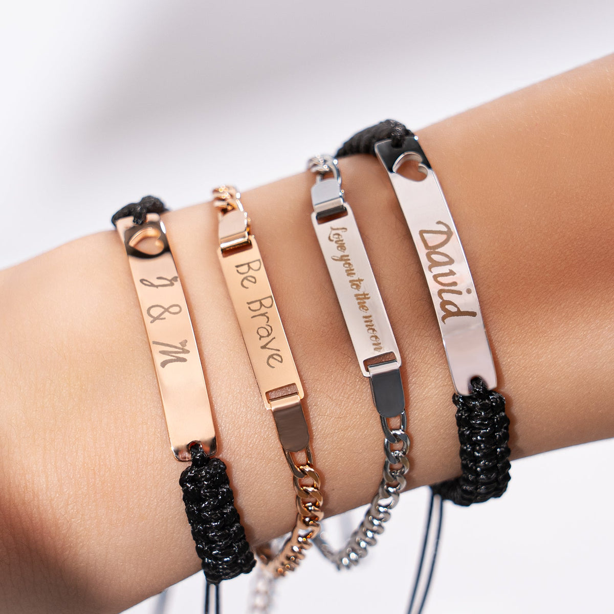 A set of personalized laser-engraved stainless steel bracelets worn layered, featuring custom engravings including initials "M & Y", the phrase "Be Brave", "I love you to the moon", and the name "David", showcasing the versatility of stainless steel bracelet blanks for laser engraving.