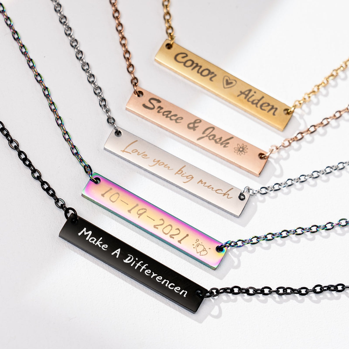 A collection of laser-engraved stainless steel bar necklaces in five finishes (black, rainbow, silver, rose gold, and gold), featuring personalized engravings such as "Make A Differencen", the date "10-19-2021", "Love you big much", "Srace & Josh", and "Conor Aiden", demonstrating the creative customization possible with laser engraving on jewelry blanks.