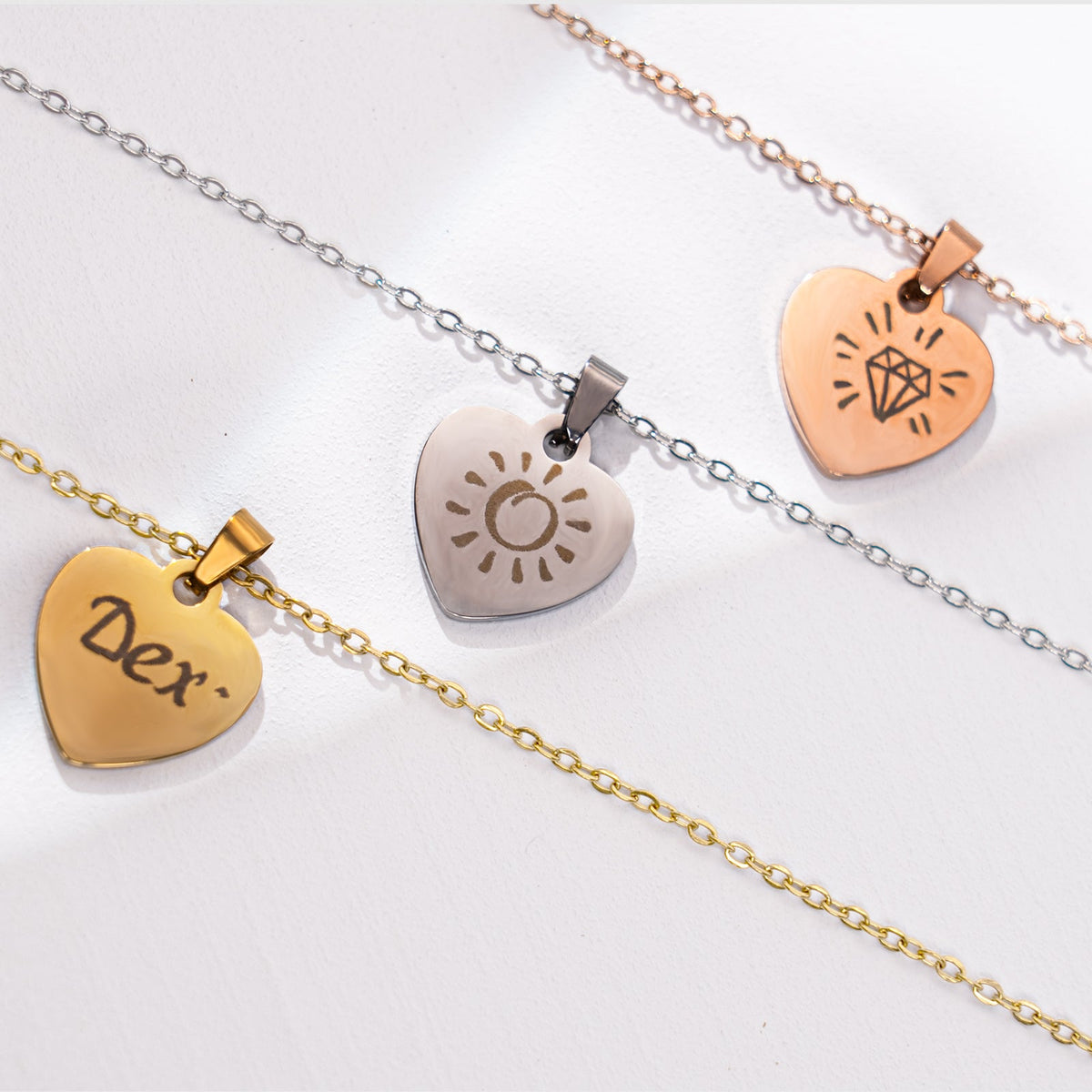 Three personalized laser-engraved heart-shaped pendant necklaces in gold, silver, and rose gold finishes, featuring the name "Dex", a sun pattern, and a diamond design, demonstrating the custom engraving possibilities of stainless steel jewelry blanks.