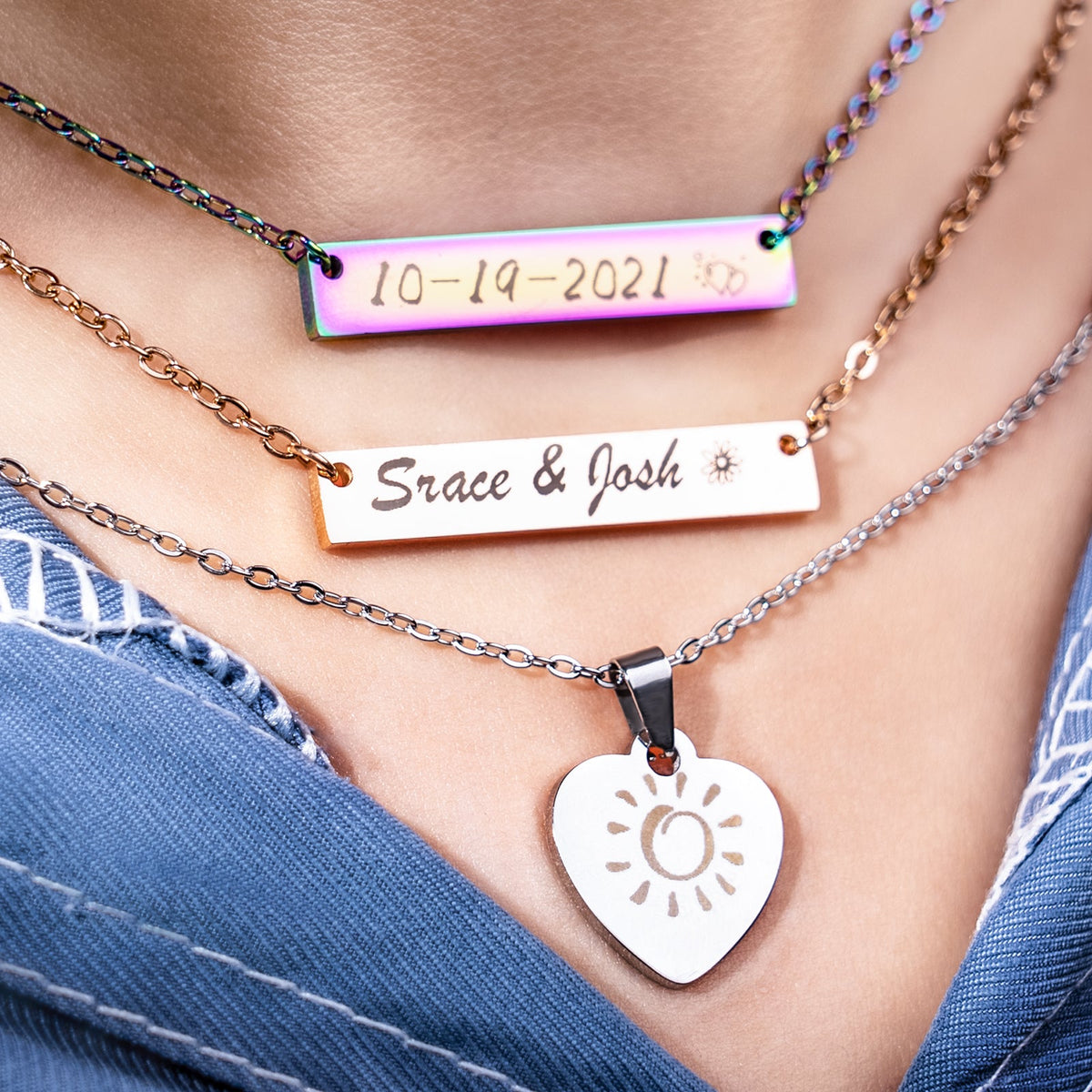 A set of personalized laser-engraved stainless steel necklaces worn layered, including a rainbow bar engraved with "10-19-2021", a classic bar engraved with "Srace & Josh", and a heart-shaped pendant with a sun pattern, demonstrating the creative customization possible with laser engraving jewelry blanks.