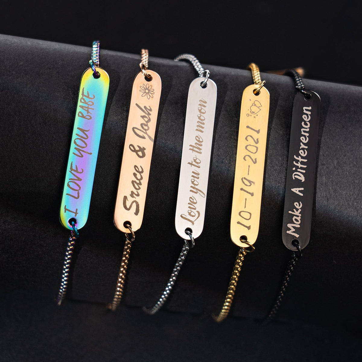 A set of five laser-engraved stainless steel bar bracelets in rainbow, rose gold, silver, gold, and black finishes, featuring personalized engravings including "I LOVE YOU BABE", "Srace & Josh", "Love you to the moon", the date "10-19-2021", and "Make A Differencen", showcasing the creative customization possible with laser engraving on jewelry blanks.