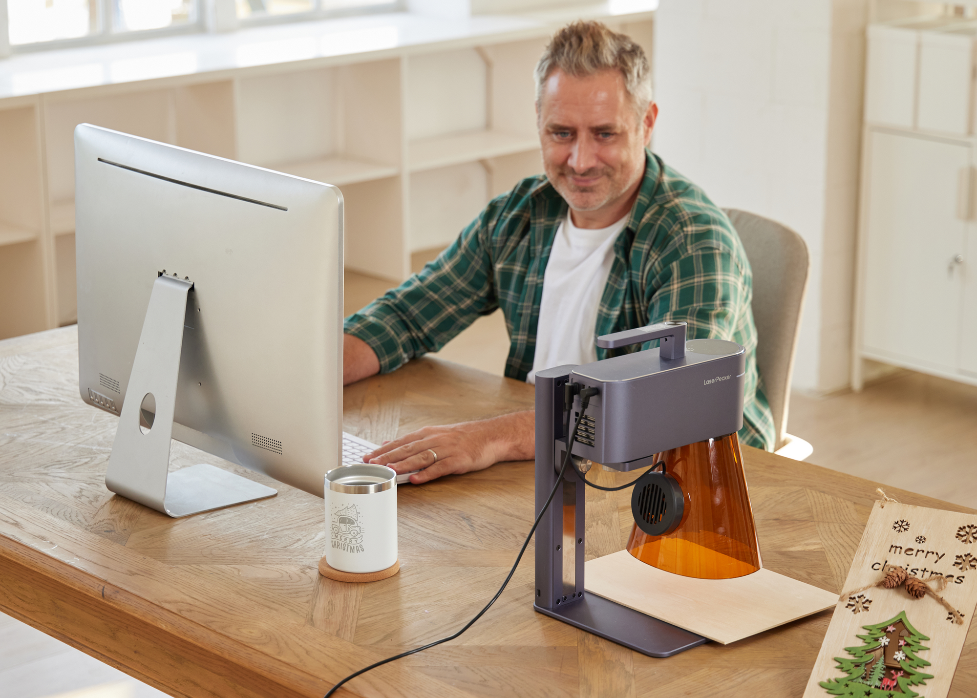A small business owner using a computer for custom product design while a LaserPecker LP4 engraves stock nearby in a workshop.