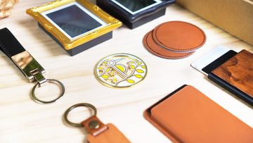 A close-up flat lay of customizable gift accessories on a light wooden surface, including ornate gold and black picture frames, a gold enamel pin with a stylized bird and foliage design, leather coasters, a black leather keychain, a wooden keychain, a tan leather cardholder, and a wooden cardholder, all designed for laser engraving personalization.