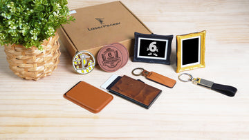 A flat lay display of LaserPecker branded customizable gift items arranged on a light wooden surface, featuring a kraft paper gift box, a gold enamel pin with a stylized bird design, a leather anniversary coaster engraved with "Celebration 6 Years Anniversary 2017-2023", an ornate black picture frame with a "6" graphic, an ornate gold picture frame, a wooden keychain, a black leather keychain, a tan leather cardholder, and a wooden cardholder, accented by a small potted plant.