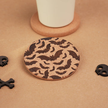 Laser engraved cork coaster with a repeating bat pattern, placed with a white mug and skull accessories.