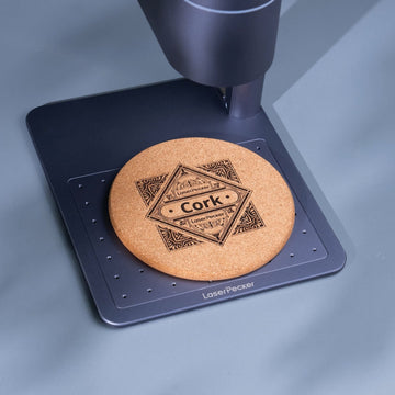 LaserPecker handheld engraver creating a geometric and text design on a cork coaster on the positioning plate.
