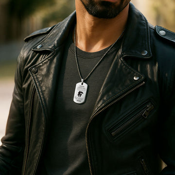 A man wearing a black leather jacket and dark t-shirt, showcasing a personalized stainless steel dog tag necklace with a beaded ball chain. The tag features custom engraved text and a silhouette design, styled for a modern, masculine look.
