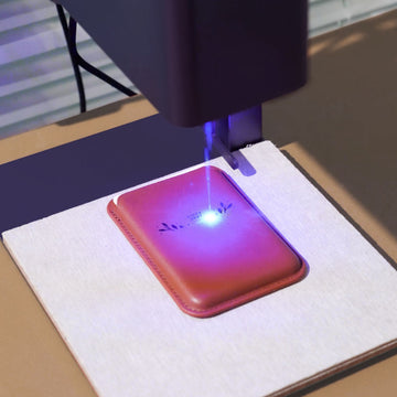 laser engraving in action, its blue laser beam actively etching a delicate design onto a brown faux leather card holder. The precision of the laser creates crisp, permanent details, showcasing how functional accessories can be transformed into personalized gifts or branded items with custom text, logos, or patterns.